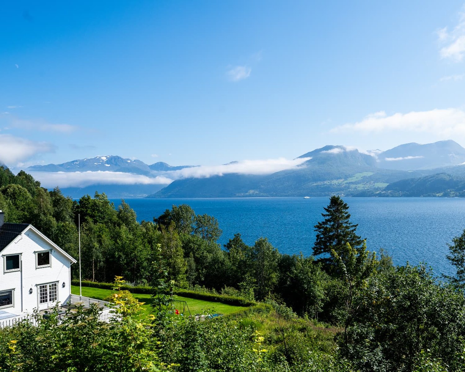 Vit trävilla med tomt och altan sedd uppifrån, med fjord och berg i bakgrunden